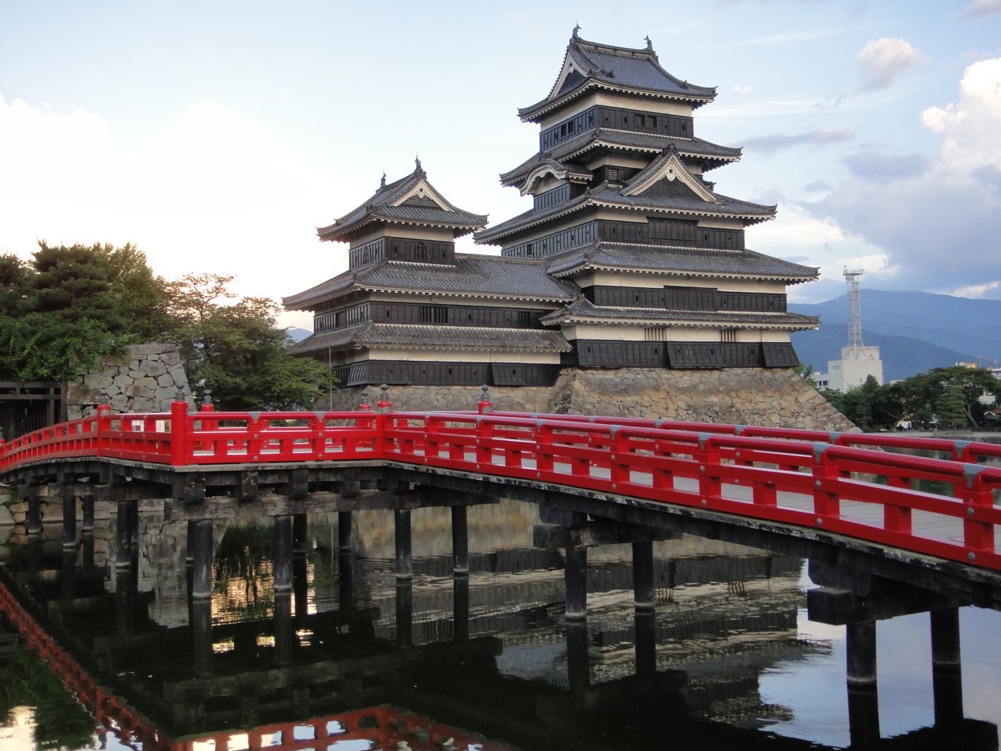 A traditional Japanese castle with multiple tiers and a distinct roof design is surrounded by water, featuring a red wooden bridge in the foreground. The scene includes lush greenery and mountains in the background, creating a serene atmosphere.