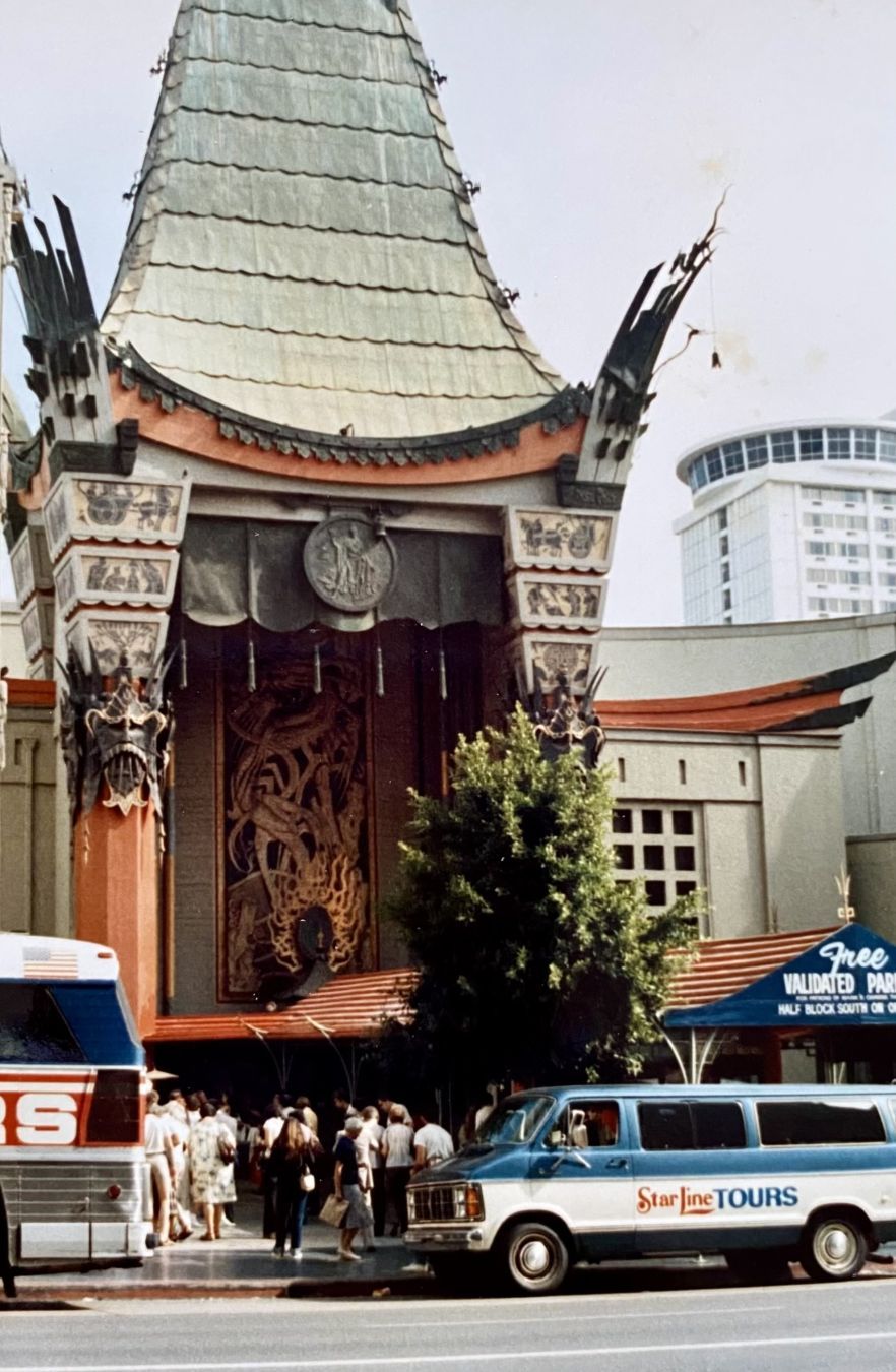 The image shows the iconic Grauman's Chinese Theatre in Hollywood, featuring its distinct architectural style and decorative elements. A crowd is gathered at the entrance, with tour vans parked nearby, including one marked 