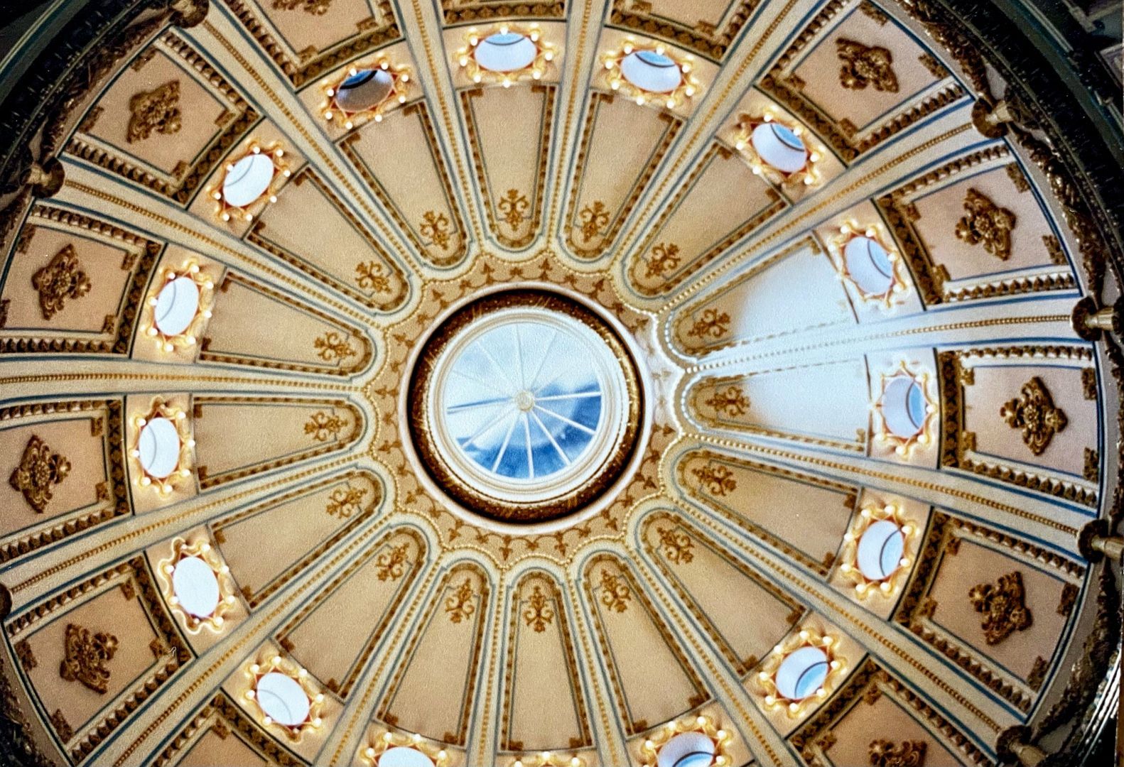 A beautifully ornate ceiling featuring a circular design with decorative patterns, small windows, and lights, viewed from below.