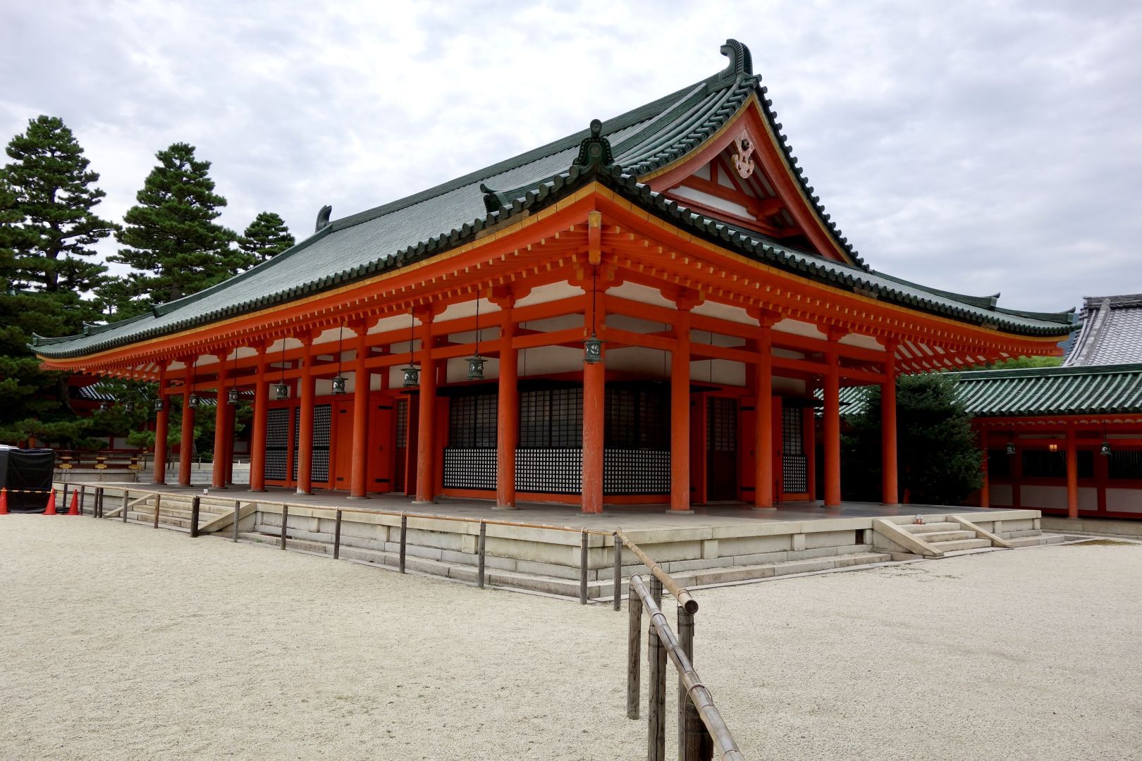 A traditional Japanese building featuring a distinctive roof and orange pillars. The structure is surrounded by a gravel area and trees, with a cloudy sky in the background.