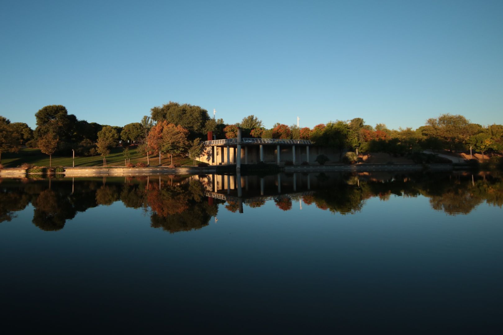 Wide angle picture of a park in front of a large body of water. The picture is cut in half by the shore, and as the water is perfectly calm it reflects the trees and structures on the other side as a mirror.