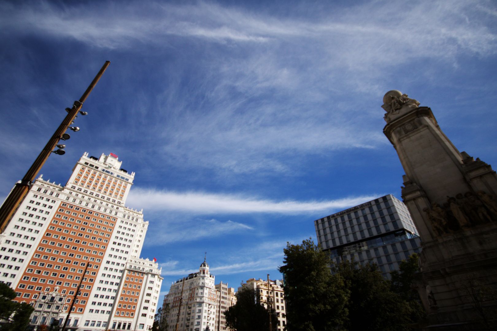 Wide angle picture from Plaza España, in Madrid. The iconic marble pillar, with people's statues and a ball on top, is on the left, in the shadow. On the right a large pole is in front, and in the back the main Plaza España hotel is found, a typical building front in orange and white. Blue sky occupies most of the picture, with light clouds throughout.