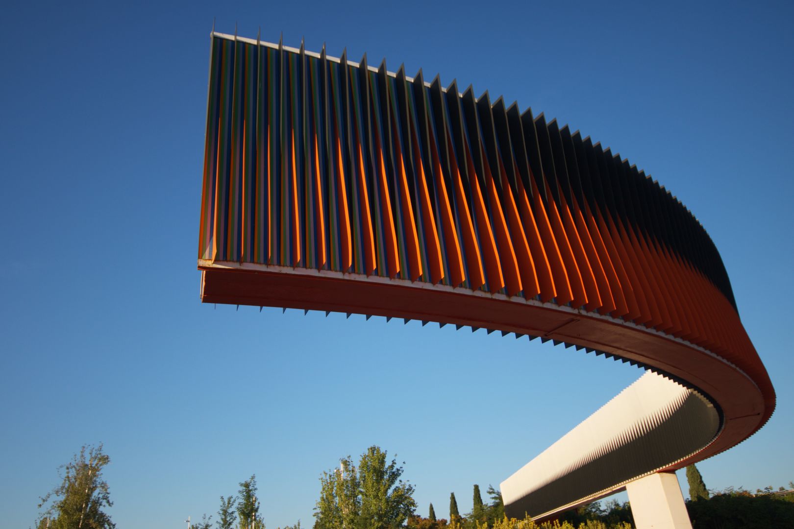 Sculpture in a park. It consists of a long, segmented band with fin-like appendages. The colors are cut in half longitudinally, on one side black on top and orange on bottom, on the other white on top and black on bottom. The band follows a very steep curve, and is held up by a pole above ground. The picture is wide angle, and the whole sculpture can be seen, from both sides due to the curve. On the bottom of the picture, trees can be seen, while the rest of the background is a bright blue sky. This was really hard to describe, I hope it came accross somewhat clear.