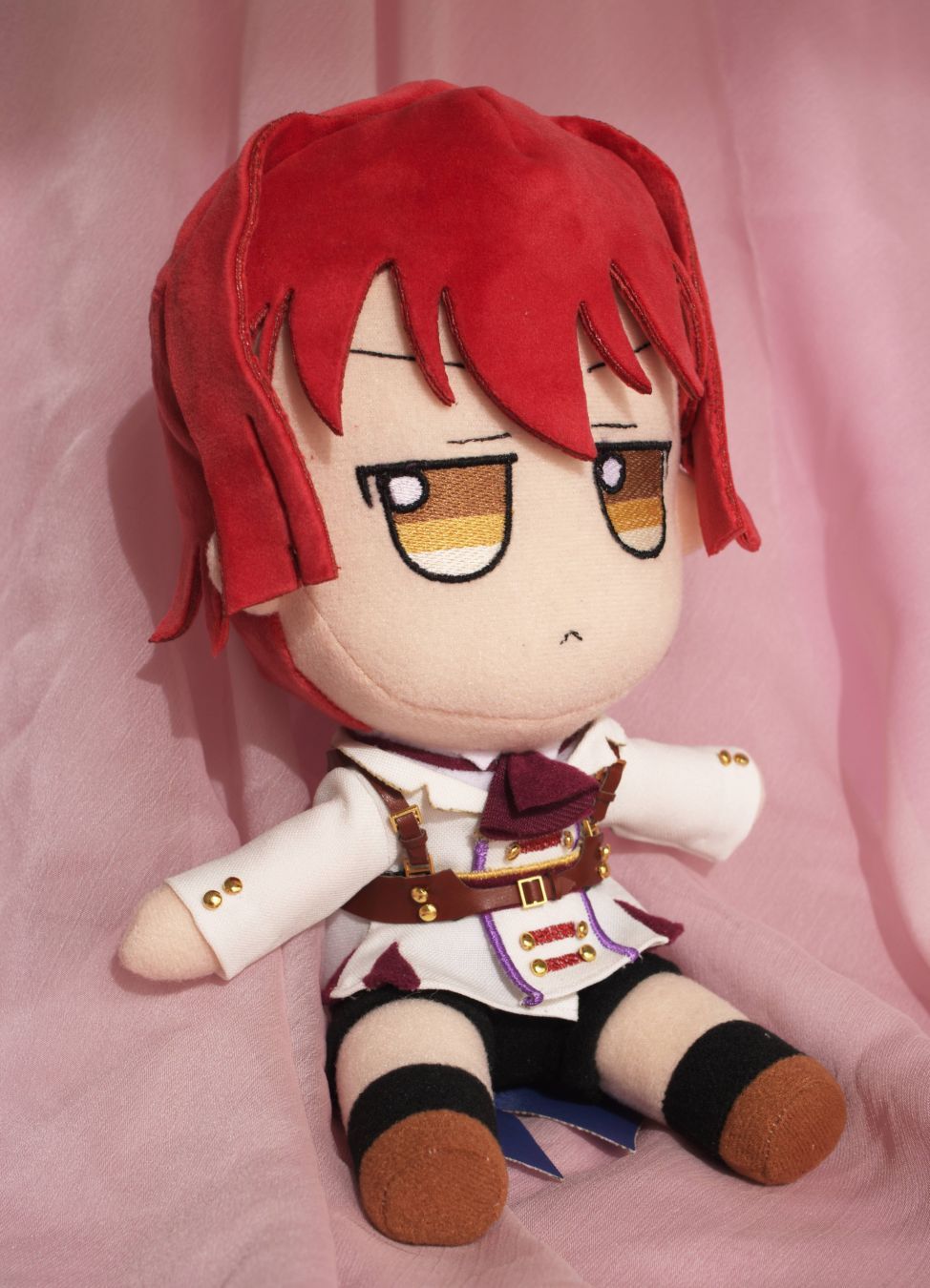 A plush, specifically a fumo plush, on a pink textile. It's Miyao, a character from the Ciconia When They Cry visual novel. It's quite stylized, with a very big head and hazel color gradient eyes, with a neutral expression. The hair is bright red, and it sports a very detailed outfit reflecting the one he wears in the novel, with a burgundy scarf on the neck, slacks and a white, somewhat military themed overall with several buttons