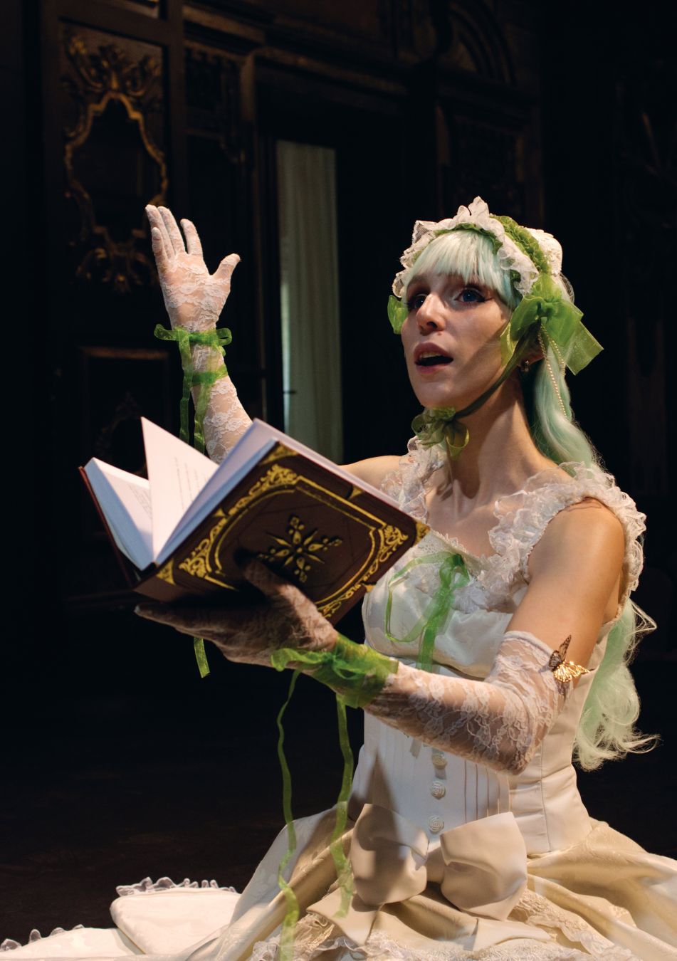 Campurrianas, the cosplayer, holding an elaborate, handmade book, with the other hand up and mouth open, as if preaching. They are portraying a young woman with long, green hair and a very ornate white dress and matching translucent and embroidered gloves. The hair is held by an embroidered tiara. It's a character from the visual novel Umineko When They Cry. The background depicts an old theater, blurred by depth of field.